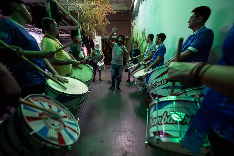 Um grupo de pessoas está ensaiando com surdos (tambores) em um ambiente interno. No centro, um homem está orientando os outros, que seguram tambores de diferentes tamanhos. A iluminação é suave e verde, e ao fundo há uma parede decorada com uma pintura. Os participantes estão vestidos com camisetas coloridas, e alguns têm baquetas nas mãos.