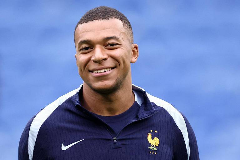 Um jovem jogador de futebol está sorrindo para a câmera. Ele usa um uniforme de treino azul escuro com detalhes brancos e um emblema que apresenta um galo, representando a seleção francesa. O fundo é desfocado em um tom de azul claro.
