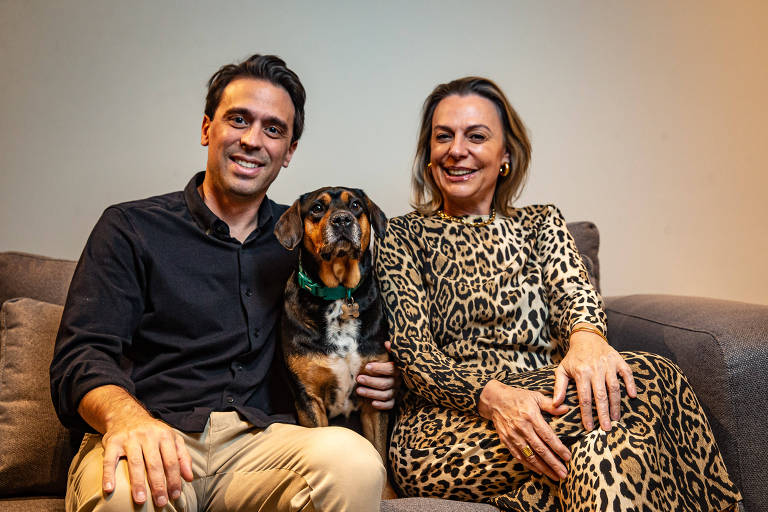 A imagem mostra um homem e uma mulher sentados em um sofá. O homem está à esquerda, vestindo uma camisa preta e calças claras, enquanto a mulher está à direita, usando um vestido com estampa de onça. Entre eles, há um cachorro de pelagem marrom e preta. O fundo é neutro e a iluminação é suave.