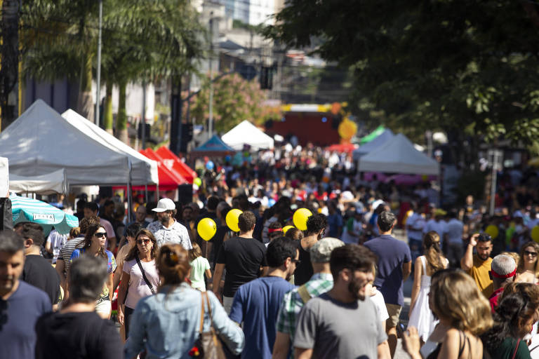 A imagem mostra uma rua movimentada durante uma feira ao ar livre, com muitas pessoas caminhando entre barracas brancas e coloridas. Ao fundo, há árvores e uma grande quantidade de pessoas, algumas usando balões amarelos. O ambiente é ensolarado e vibrante.