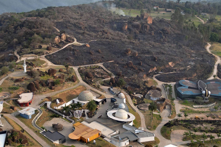 Imagem aérea de uma área que foi afetada por um incêndio. O terreno apresenta grandes áreas queimadas, com vegetação destruída. No primeiro plano, há várias estruturas arquitetônicas, incluindo edifícios de formas variadas, como domos e edifícios circulares, além de estradas e caminhos visíveis. Ao fundo, é possível ver fumaça