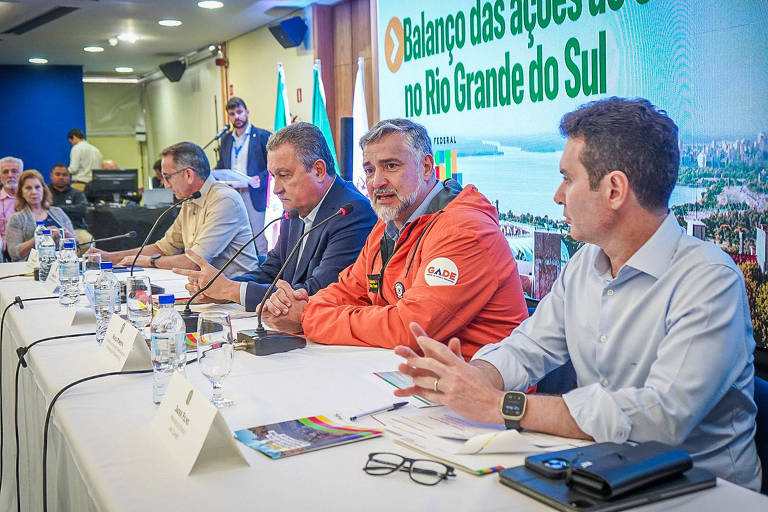 Na foto, quatro homens estão sentados à mesa, e um homem branco grisalho de jaqueta laranja fala ao microfone