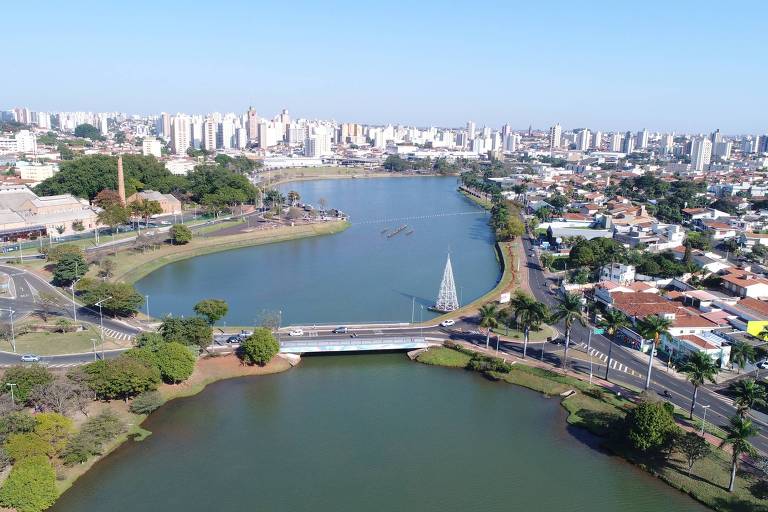 A imagem mostra uma vista aérea de São José do Rio Preto com uma represa em primeiro plano. À esquerda, há uma área verde com árvores e uma estrada que contorna a represa