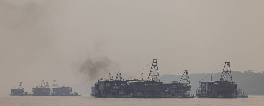 A imagem mostra vários navios ancorados em uma área aquática, com fumaça saindo de um deles. O ambiente é nebuloso, dificultando a visibilidade, e os navios possuem estruturas de guindaste visíveis.