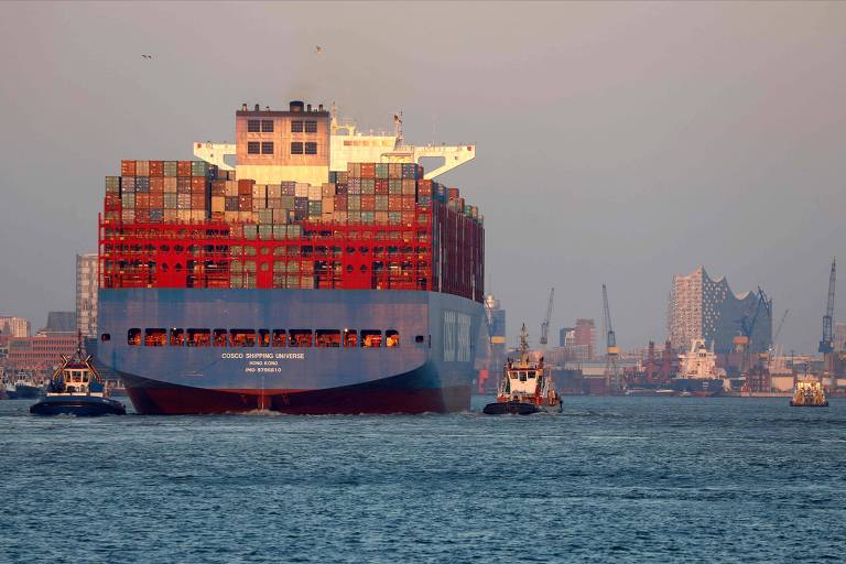 A imagem mostra um grande navio de carga, carregado com contêineres coloridos, navegando em um porto. O navio é de cor azul na parte inferior e vermelho na parte superior, com várias janelas visíveis. Há também barcos de apoio ao redor do navio. Ao fundo, é possível ver a silhueta de edifícios e estruturas portuárias, incluindo um edifício moderno com uma forma distinta.