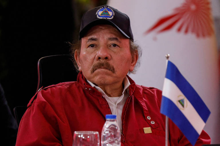 Daniel Ortega está sentado, usando um boné preto e uma jaqueta vermelha. Ele tem uma expressão séria e está olhando para frente. À sua frente, há uma garrafa de água e uma pequena bandeira da Nicarágua