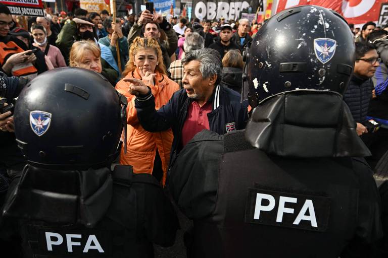 A imagem mostra um confronto entre manifestantes e policiais em um protesto. Um homem idoso, com cabelo grisalho e vestindo uma jaqueta escura, está gesticulando e falando intensamente. Ao seu lado, uma mulher com cabelo loiro e preso, vestindo um casaco laranja, observa. Dois policiais, com capacetes e coletes pretos marcados com 'PFA', estão de costas, observando a situação. Ao fundo, há uma multidão de pessoas e cartazes visíveis.
