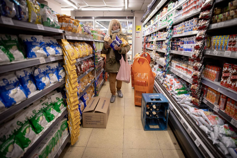 Uma mulher está em um corredor de supermercado, segurando pacotes de produtos alimentícios. Ela usa um casaco e uma máscara, e tem cabelo loiro. Ao seu redor, prateleiras estão cheias de produtos embalados, como biscoitos e laticínios. No chão, há uma caixa de papelão e uma cesta de compras laranja. O ambiente é bem iluminado.
