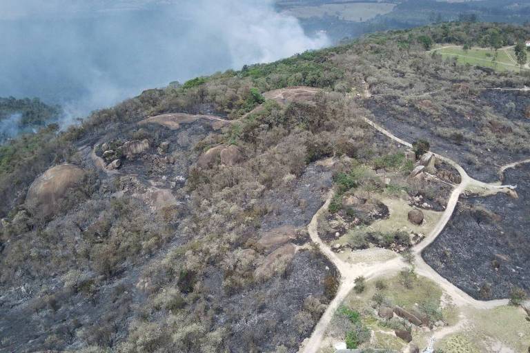 Imagem aérea de uma área afetada por incêndio florestal, mostrando terrenos queimados e vegetação danificada. O solo está coberto por cinzas e restos de plantas, com algumas trilhas visíveis que cortam a paisagem. Ao fundo, há fumaça se elevando, indicando a presença de fogo nas proximidades