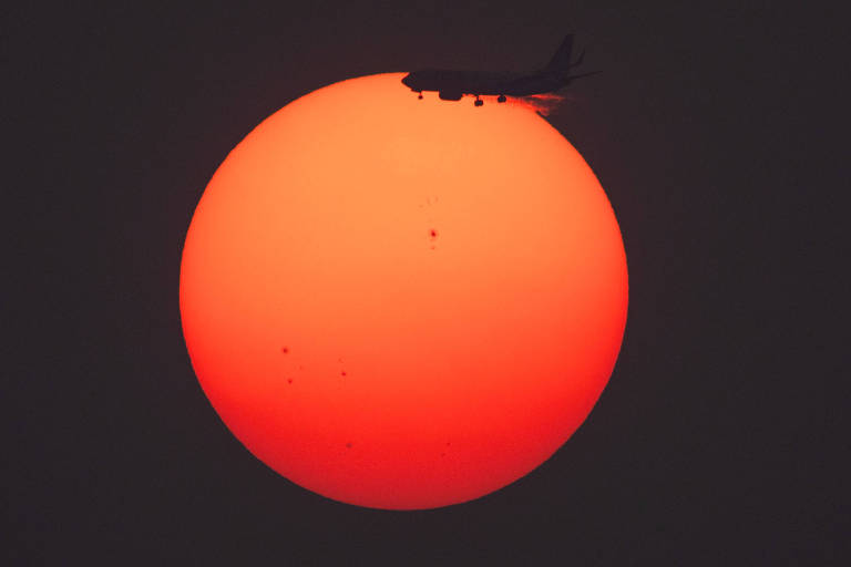 A imagem mostra um grande sol avermelhado no fundo, com um avião em silhueta voando na frente dele. O sol é redondo e apresenta manchas escuras em sua superfície, enquanto o avião é visível apenas como uma sombra.