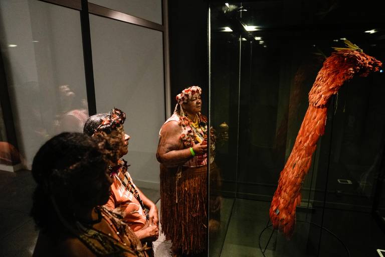 A imagem mostra três mulheres indígenas observando um objeto exposto em uma vitrine. As mulheres estão vestidas com trajes tradicionais, adornadas com colares e tiaras feitas de flores e penas. O objeto em exibição é uma peça longa e vermelha, feita de penas, que se destaca na vitrine iluminada. O ambiente é escuro, com reflexos visíveis nas superfícies de vidro.