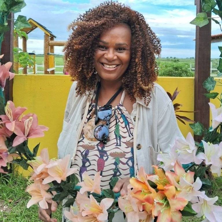 Uma mulher com cabelo cacheado e volumoso está sorrindo em um ambiente ao ar livre, cercada por flores em tons de rosa e laranja. Ela usa uma blusa colorida e um casaco claro. Ao fundo, há uma estrutura amarela e um céu azul com nuvens.