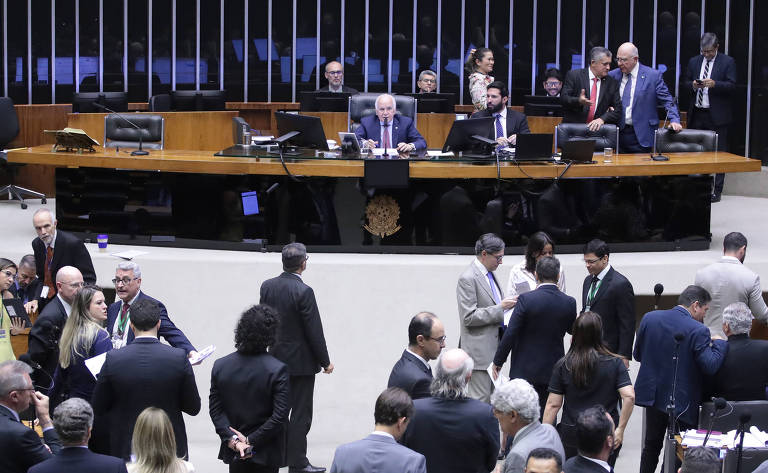 A imagem mostra uma sessão na Câmara dos Deputados do Brasil, com vários deputados em pé e sentados ao redor de uma mesa central. Um deputado está falando ao microfone, enquanto outros discutem entre si. O ambiente é formal, com computadores e documentos visíveis. Ao fundo, há uma série de cadeiras e mesas organizadas.