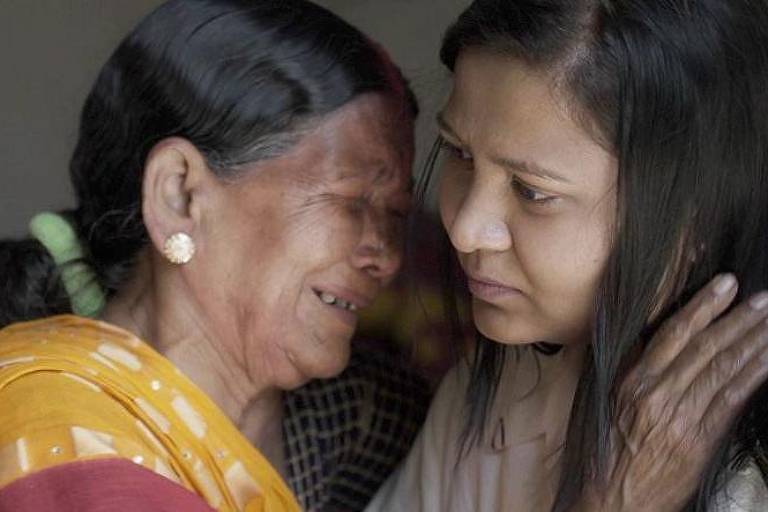 A imagem mostra duas mulheres em um momento emocional. A mulher mais velha, com cabelo preto e liso, está chorando e parece estar consolada pela mulher mais jovem ao seu lado. A jovem, com cabelo longo e liso, olha para a mulher mais velha com uma expressão de empatia. Ambas estão em um ambiente interno, com uma iluminação suave.
