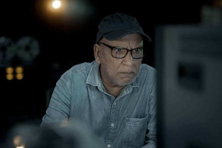 Um homem idoso, usando óculos e um boné, está concentrado em frente a um computador. Ele veste uma camisa jeans e parece estar em um ambiente com pouca luz, onde há algumas luzes ao fundo.
