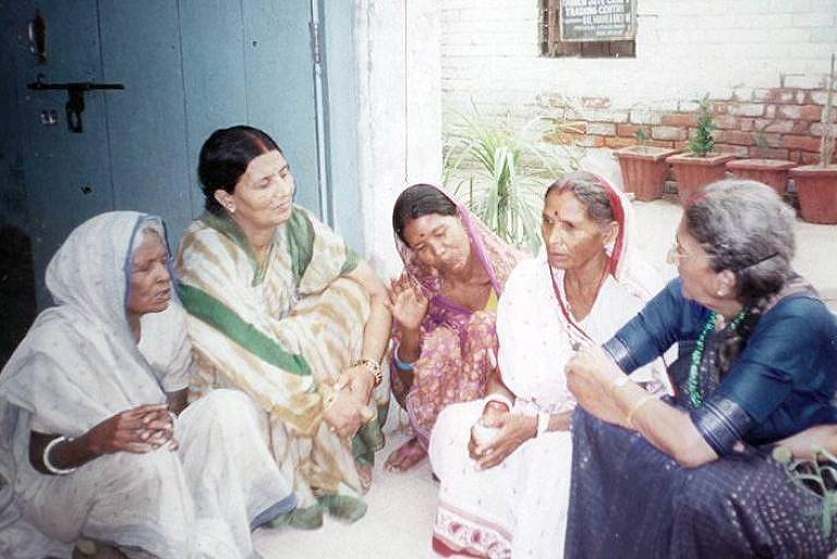 A imagem mostra um grupo de cinco mulheres sentadas em um ambiente externo, próximo a uma porta azul. Elas estão vestidas com roupas tradicionais e parecem estar envolvidas em uma conversa. O fundo apresenta um espaço com plantas e um ambiente residencial.
