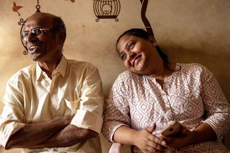 A imagem mostra um homem idoso e uma mulher sentados lado a lado, ambos sorrindo. O homem usa óculos e uma camisa clara, enquanto a mulher veste uma blusa de padrão claro. O fundo é de uma parede simples, com uma iluminação suave que destaca os rostos deles.
