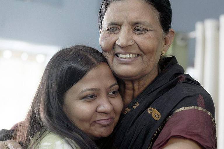 A imagem mostra duas mulheres se abraçando. A mulher à esquerda tem cabelo longo e liso, e está sorrindo. A mulher à direita tem cabelo curto e está usando uma roupa tradicional. Ambas parecem felizes e estão em um ambiente interno com paredes claras.
