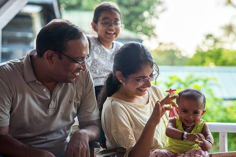 A imagem mostra uma família sentada em uma varanda. Um homem e uma mulher estão à frente, ambos sorrindo e interagindo com uma criança pequena que está no colo da mulher. Ao fundo, uma menina está sorrindo e observando. A cena é iluminada pela luz natural, com vegetação visível ao fundo.
