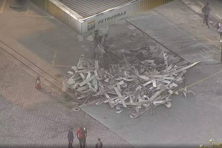 A imagem mostra os destroços de uma estrutura desabada em um posto de combustível da Petrobras. Há uma pilha de materiais, como placas de concreto e metal, acumulados no chão. Algumas pessoas estão próximas ao local, observando a situação. O ambiente é urbano, com pavimentação de paralelepípedos e uma estrutura de posto visível ao fundo.