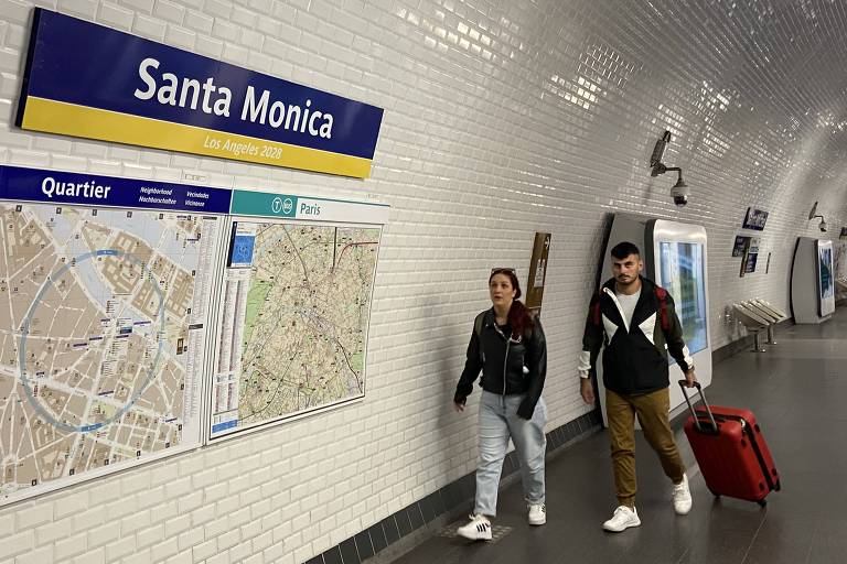 A imagem mostra a estação de metrô Santa Monica, com um painel azul indicando o nome da estação. À esquerda, há um mapa da área. Duas pessoas estão caminhando na plataforma, uma delas puxando uma mala vermelha. O ambiente é iluminado e as paredes são revestidas de azulejos brancos.