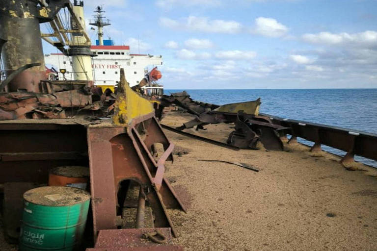 Imagem mostra convés de navio, com a torre de comando ao fundo, com danos. Há metal retorcido em vários pontos. O mar está ao fundo, com um céu azul com nuvens