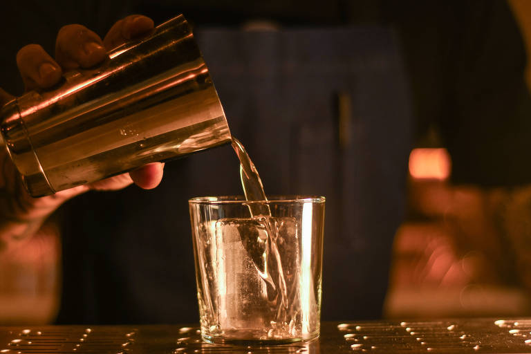 Um bartender está despejando um líquido de um shaker metálico em um copo de vidro. O fundo é iluminado com uma luz suave.