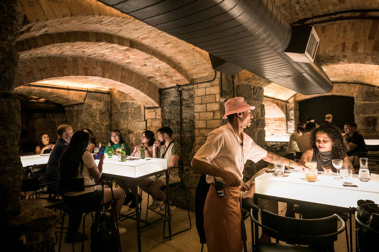 A imagem mostra um bar localizado em um ambiente subterrâneo, com paredes de pedra e arcos no teto. Há várias mesas com clientes sentados, alguns conversando e outros consumindo bebidas. Uma funcionária, vestindo uma camisa clara e um chapéu rosa, está servindo em uma das mesas. A iluminação é suave.