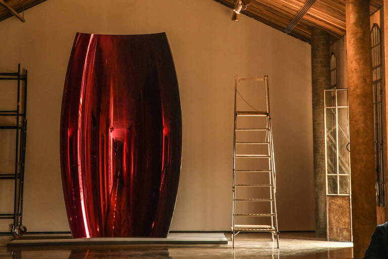 A imagem mostra um espaço interno com um teto de madeira, onde há uma grande obra de arte de cor vermelha brilhante em forma oval. À esquerda, há uma estrutura de andaime e, à direita, janelas com molduras de vidro. O piso é liso e reflete a luz. O ambiente é bem iluminado por spots de luz no teto.