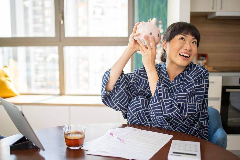 Mulher segurando cofrinho, sorrindo e com papéis e calculadora em cima da mesa
