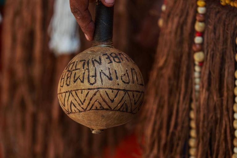 A imagem mostra uma mão segurando um maracá, instrumento tradicional, com inscrições gravadas em sua superfície. O fundo é desfocado, mas parece haver elementos decorativos, como fios e contas coloridas, sugerindo um contexto cultural. O maracá tem um padrão de linhas e formas geométricas na parte inferior.