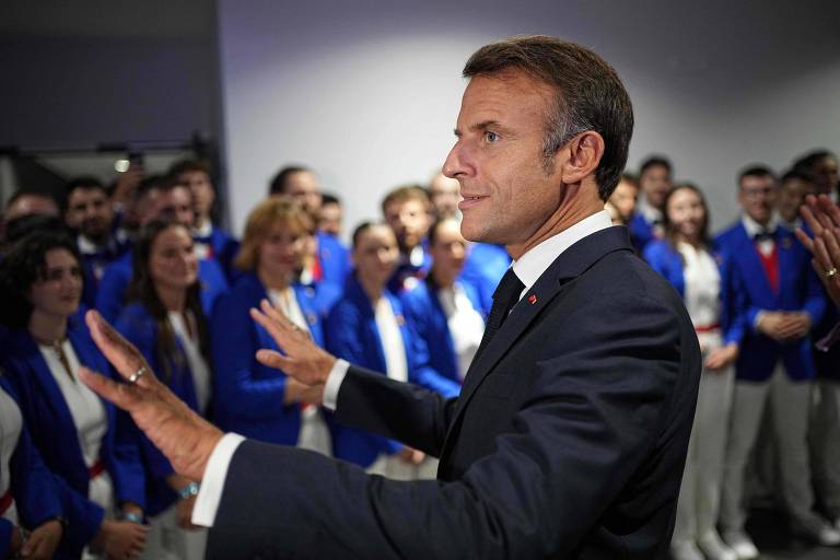 A imagem mostra Emmanuel Macron, presidente da França, um homem em um terno escuro, com cabelo curto e uma expressão amigável, gesticulando com as mãos. Ele está em primeiro plano, enquanto ao fundo há um grupo de pessoas vestindo blazers azuis e camisas brancas, sorrindo e observando. O ambiente parece ser um evento social ou político.