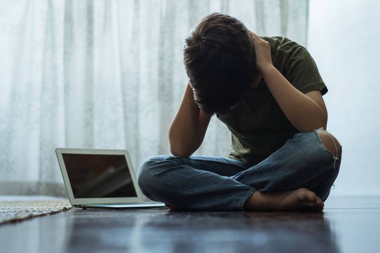 Fotografia mostra a imagem de um menino triste, sentado no chão de cabeça baixa e com as mãos no pescoço, ao lado de um computador