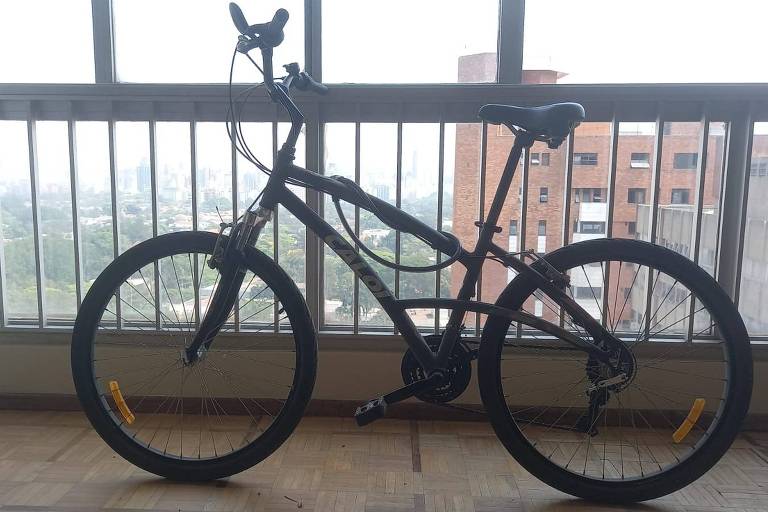 Uma bicicleta Caloi 500 aro 26 em frente a uma enorme janela de apartamento com prédios ao fundo