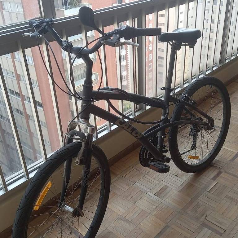 Uma bicicleta Caloi 500 aro 26 em frente a uma enorme janela de apartamento com prédios ao fundo