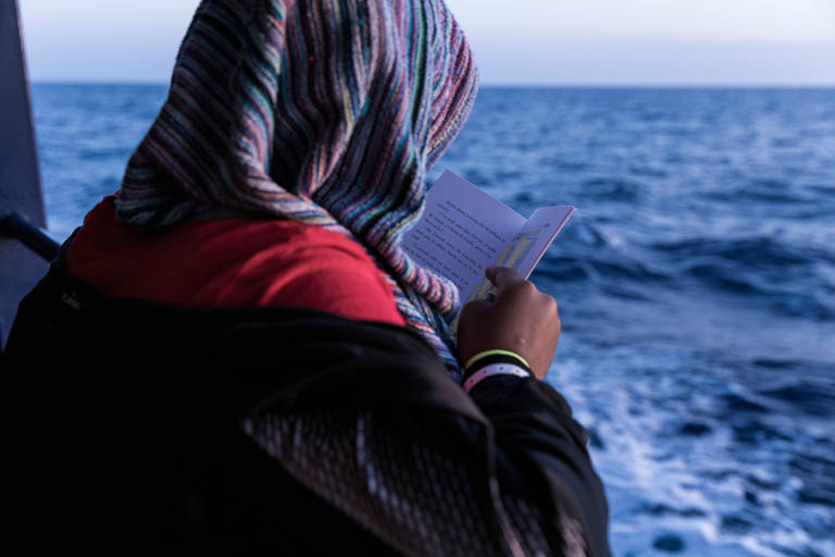 A imagem mostra uma pessoa de costas, usando um lenço colorido na cabeça, segurando um papel enquanto observa o mar. O fundo é composto por água azul e um céu claro, sugerindo um ambiente tranquilo.