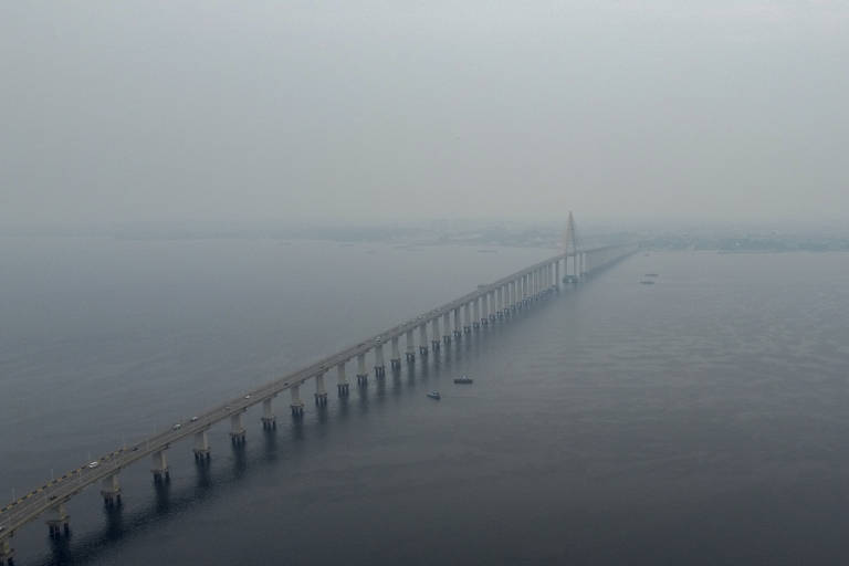 Imagem aérea de uma ponte longa que se estende sobre um corpo d'água, com um céu nublado e uma visibilidade reduzida. A ponte é composta por várias colunas que sustentam a estrutura, e o mar parece calmo, refletindo a atmosfera cinza do dia.