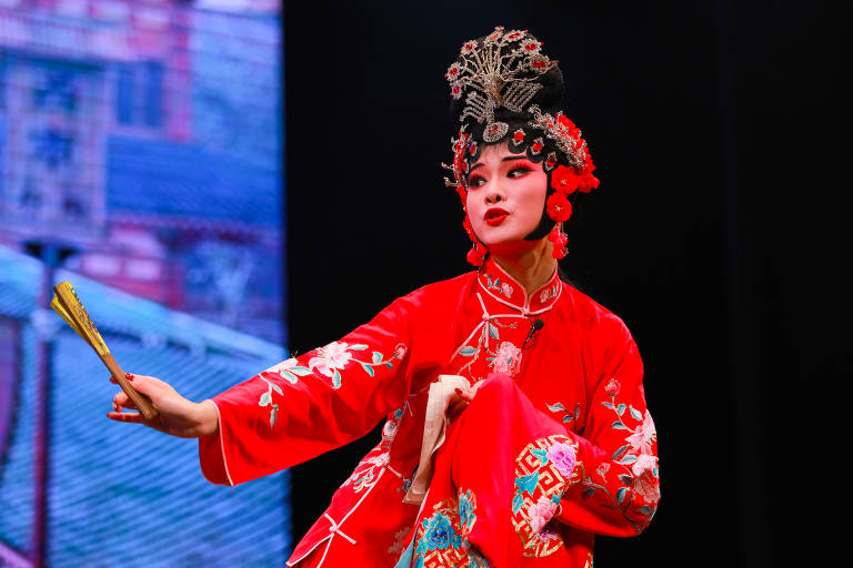 Uma artista de ópera chinesa está em cena, vestindo um traje vermelho vibrante com bordados florais. Ela segura um leque na mão esquerda e faz uma pose expressiva, com maquiagem elaborada e um penteado ornamentado. O fundo é escuro, destacando a figura da artista.