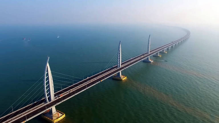 A imagem mostra uma ponte construída sobre o mar. A estrutura da ponte é longa, estendendo-se até o horizonte, com grandes pilares de sustentação em intervalos regulares. As torres de suspensão da ponte são finas e altas, com cabos de aço que se conectam ao tabuleiro, conferindo estabilidade à construção. A água ao redor parece calma, e a visão aérea dá uma perspectiva da grandiosidade da ponte, destacando sua extensão e engenharia moderna. O céu é claro e a atmosfera em torno da ponte é leve, sugerindo um dia ensolarado e condições climáticas favoráveis.