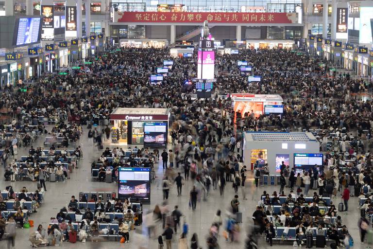 A imagem mostra o movimentado saguão de uma estação ferroviária em Xangai, na China. O espaço é vasto e repleto de pessoas, com centenas de indivíduos caminhando, aguardando ou se dirigindo aos seus destinos. No centro do saguão, há várias estruturas e quiosques iluminados, com telas exibindo informações, provavelmente sobre horários de trens e destinos. Os bancos estão lotados, e há uma sensação de grande fluxo de passageiros. O ambiente é bem iluminado e moderno, com placas de sinalização em chinês espalhadas pelo local. A quantidade de pessoas sugere um momento de grande movimentação, como em um horário de pico ou período de férias.