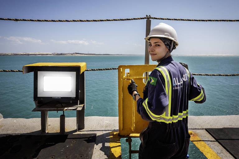 Uma mulher vestindo um uniforme de trabalho e um capacete de segurança está de pé em um cais, olhando para trás. Ao fundo, há um monitor de controle com a tela em branco e um painel amarelo ao lado. O mar é visível atrás dela, com um céu claro.
