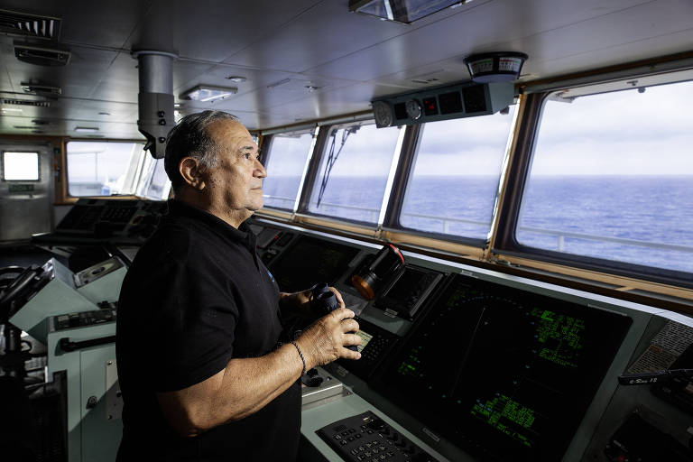 Um homem está em pé no convés de um navio, olhando pela janela em direção ao mar. Ele segura um objeto em suas mãos e está vestido com uma camisa preta. O ambiente é moderno, com painéis de controle visíveis ao fundo e janelas amplas que mostram o oceano.
