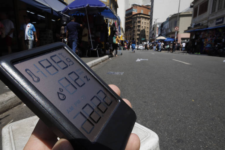 Repórter segura um termo-higrômetro, que mostra a temperatura e a umidade do ar; ao fundo, vista do asfalto sob sol quente