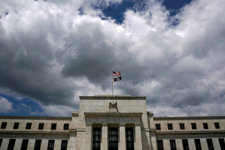 A imagem mostra a fachada do edifício do Federal Reserve, com nuvens escuras e um céu parcialmente nublado ao fundo. No topo do edifício, há duas bandeiras: a bandeira dos Estados Unidos e uma bandeira do estado. A estrutura é feita de pedra clara e possui janelas retangulares.
