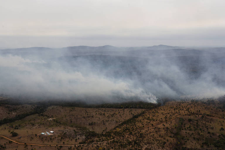 A imagem mostra uma vista aérea de uma área rural afetada por um incêndio florestal. Há fumaça densa se espalhando pela vegetação, com algumas áreas queimadas visíveis. No fundo, montanhas se estendem sob um céu nublado, enquanto uma pequena construção branca é visível no canto inferior esquerdo.
