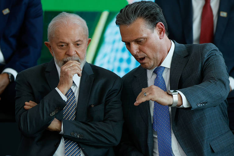 A imagem mostra dois homens sentados em um evento. O homem à esquerda, com cabelo grisalho e barba, está pensativo, com a mão no queixo. O homem à direita, com cabelo escuro e bem vestido, está gesticulando enquanto fala. Ao fundo, outras pessoas estão parcialmente visíveis, mas não são claramente identificáveis.
