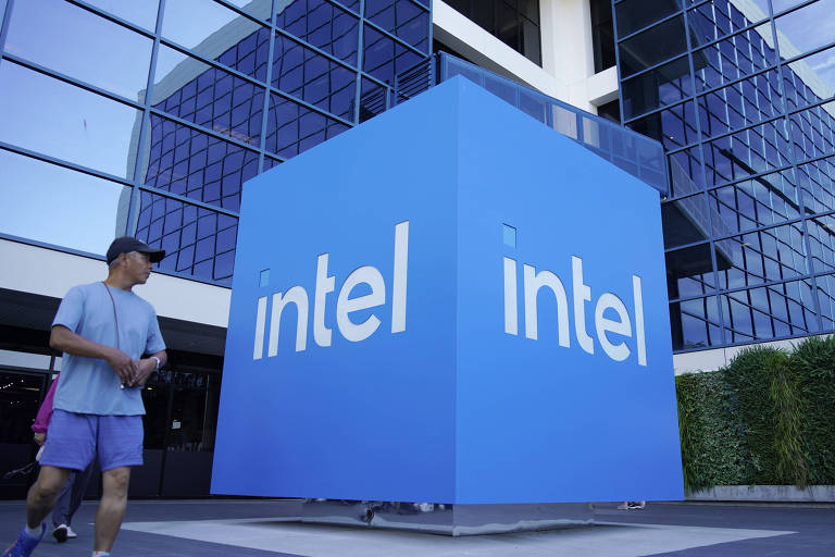 A imagem mostra a fachada de um edifício da Intel, com um grande bloco azul exibindo o nome 'intel' em letras brancas. Um homem está caminhando em frente ao bloco, e ao fundo, há janelas reflexivas do edifício. A vegetação está presente ao lado direito da imagem.
