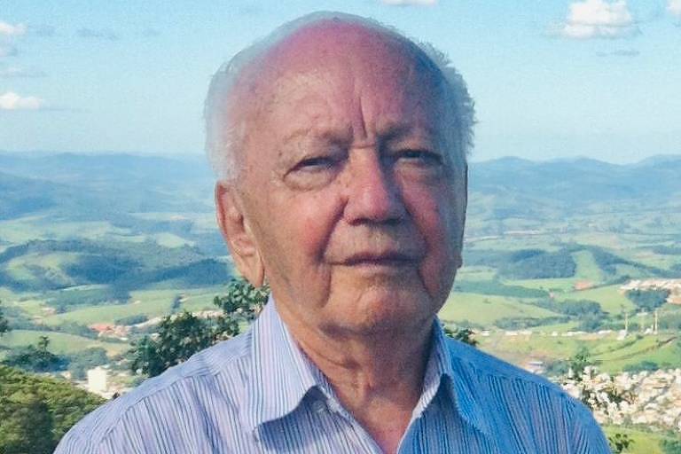 Um homem idoso está posando para a foto em uma paisagem montanhosa. Ele usa uma camisa de botão listrada em azul e branco e está em pé, com um fundo de montanhas verdes e céu azul. O homem tem cabelo grisalho e uma expressão séria.