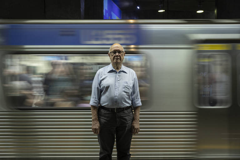 Um homem calvo e idoso está parado em uma estação de metrô, enquanto um trem passa rapidamente ao fundo, criando um efeito de movimento. O homem usa uma camisa clara e calças escuras, e parece estar em foco, enquanto o trem é borrado devido à velocidade.