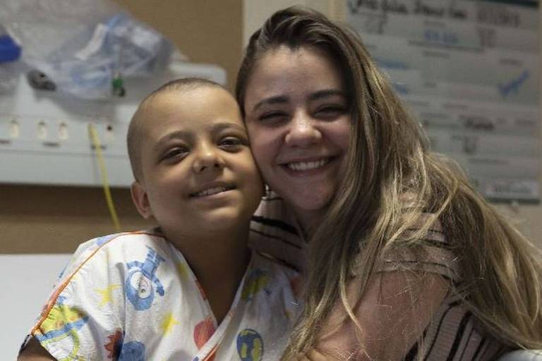 A imagem mostra uma criança com cabelo raspado, vestindo uma camisa de hospital, sorrindo e abraçando uma mulher ao seu lado. A mulher tem cabelo longo e solto, e também sorri. O fundo é um ambiente hospitalar, com paredes claras e alguns cartazes visíveis.